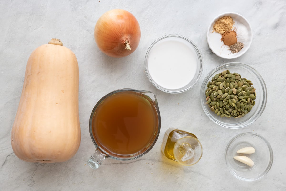 Ingredients for recipe before being prepped: butternut squash, onion, broth, cream, spices, oil, pumpkin seeds, and garlic cloves.