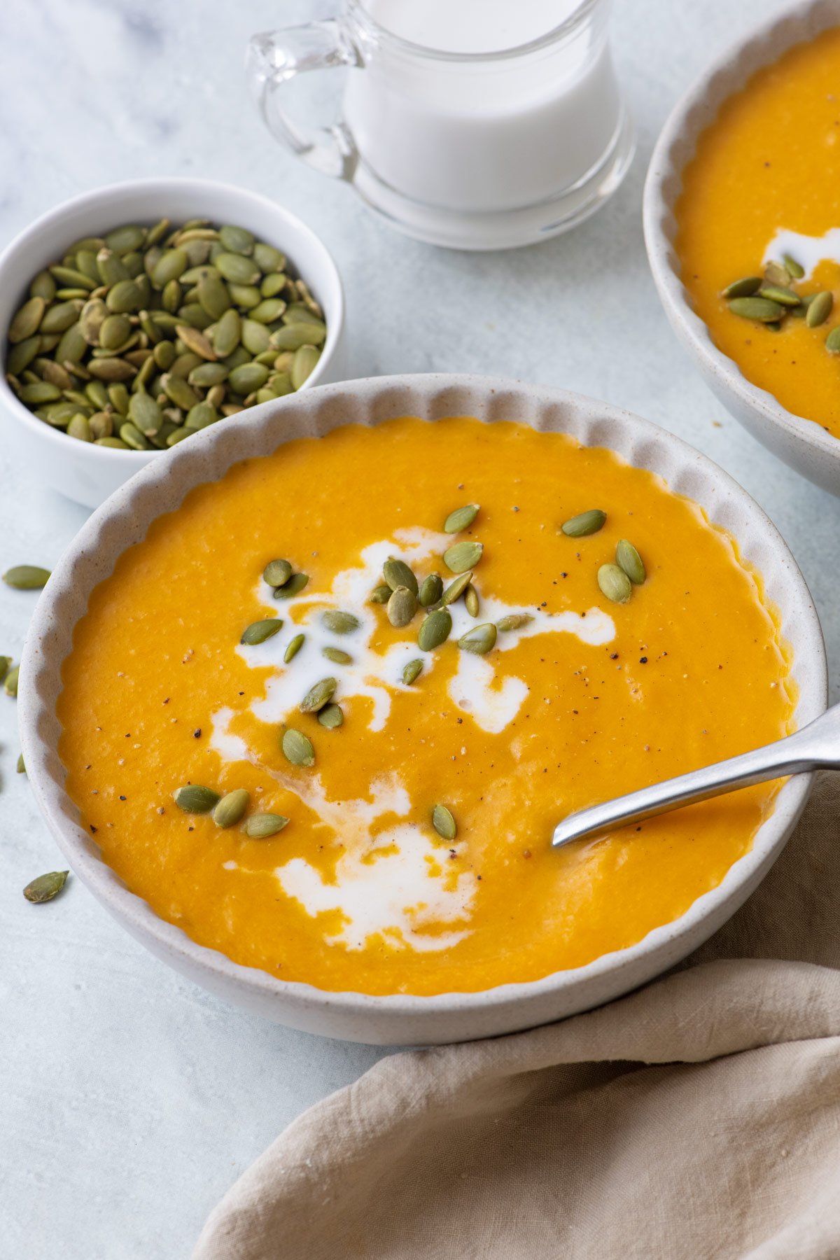 Large bowl of butternut squash soup with cream drizzled in and topped with pumpkin seeds with more nearby.