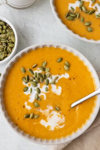 Bowl of soup garnished with coconut milk and pumpkin seeds with spoon inside bowl and another bowl nearby.