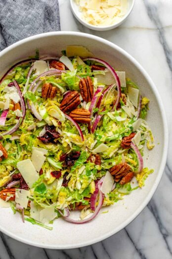 Large bowl of shaved brussel sprouts salad topped with pecans, onions, cheese, cranberries and red onions