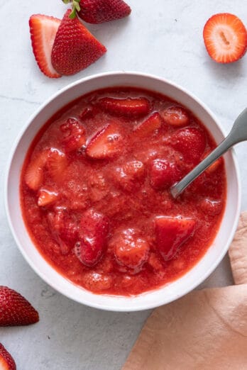 Homemade strawberry sauce in white bowl with a spoon and cut fresh strawberries around bowl.