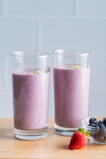 Two tall glasses of banana berry smoothies with strawberry and blueberries next to them