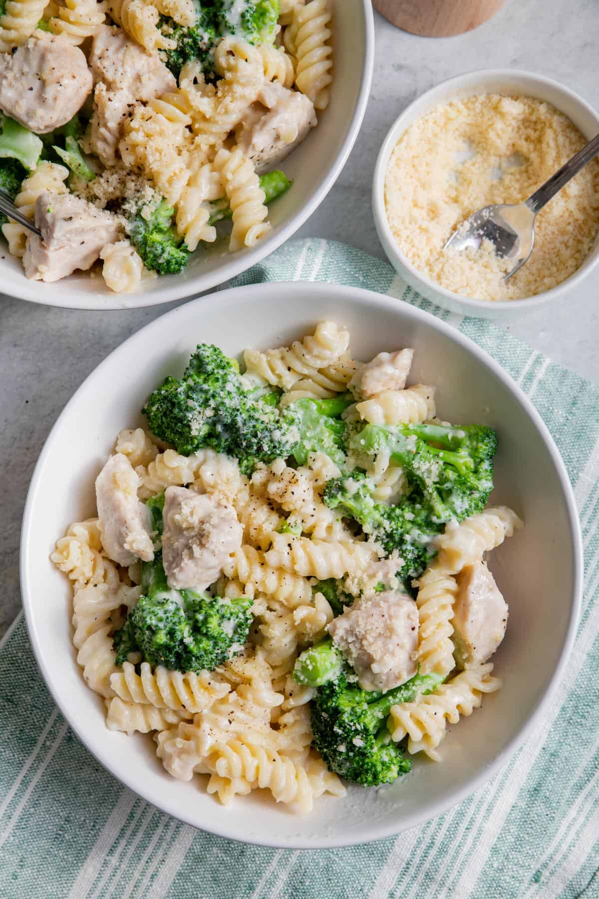 Two bowls of broccoli chicken alfredo with parmesan cheese on the side
