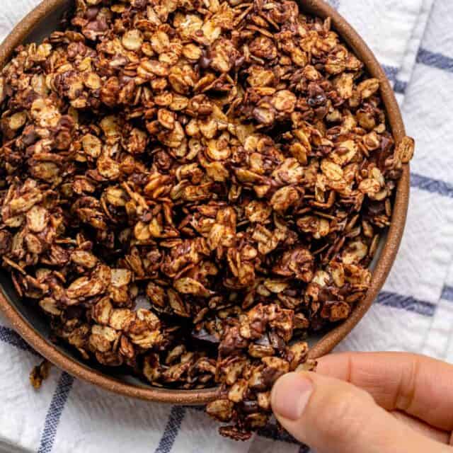 Shallow bowl of homemade chocolate granola with fingers grabbing large chunk