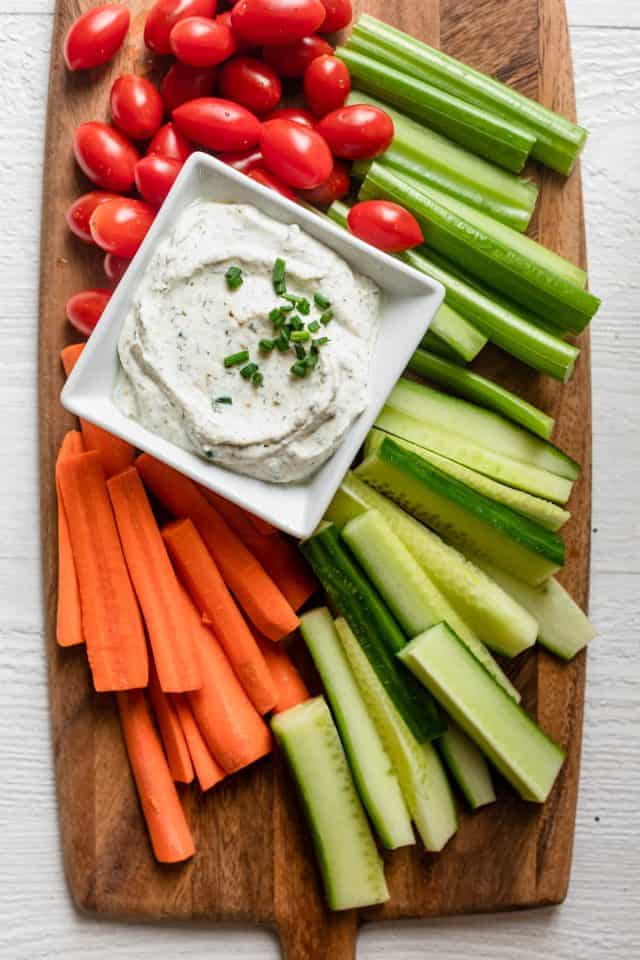 Ranch dip on wooden board surrounded by carrots, celery, cucumbers and cherry tomatoes