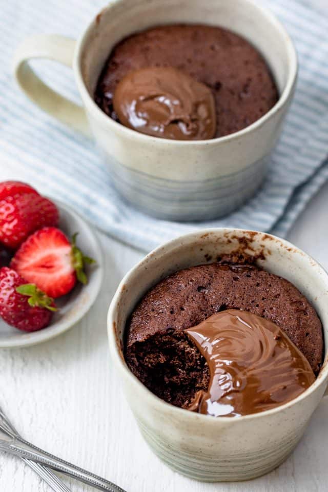 Nutella mug cake in two mugs with bite taken out of one - small plate of strawberries next to them
