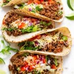 Row of ground beef tacos with colorful toppings, and limes on the side