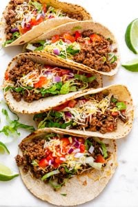 Row of ground beef tacos with colorful toppings, and limes on the side