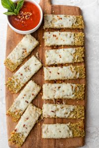 Quinoa Pizza Crust Breadsticks on a cutting board topped with crushed pepper and marinara sauce