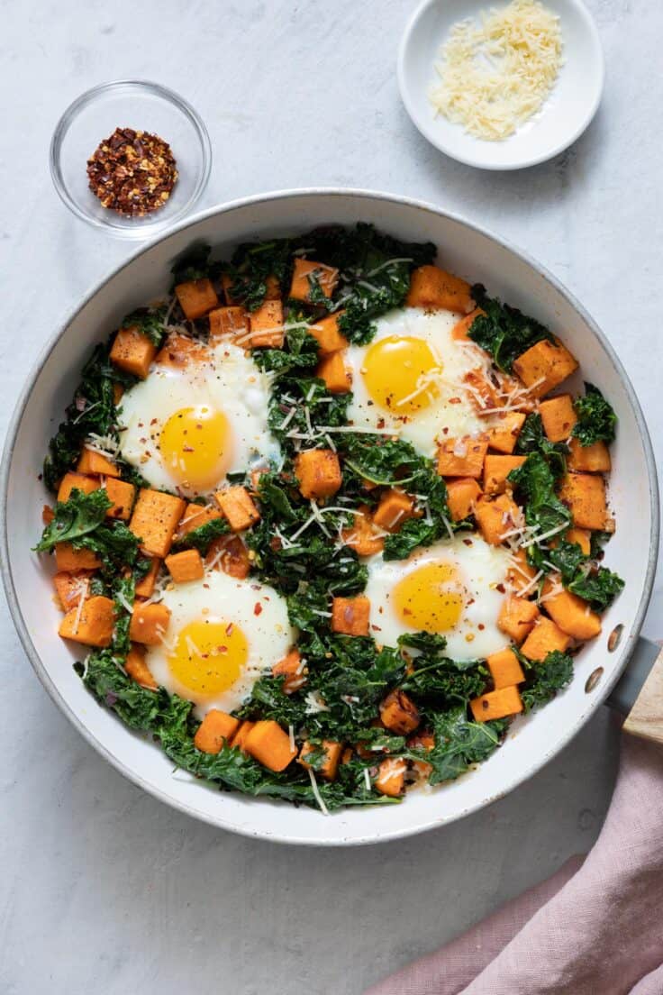 Sweet potato and kale hash with four sunny up eggs cooked in garnished with red pepper flakes and Parmesan cheese.