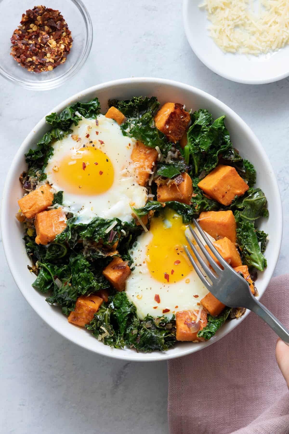 Bowl of the kale, sweet potatoes and 2 eggs and fork inside bowl, cutting into egg garnished with red pepper flakes.