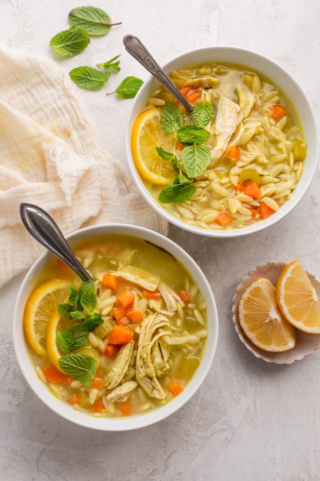 Two large bowls of chicken lemon orzo soup with slices of lemons on the side