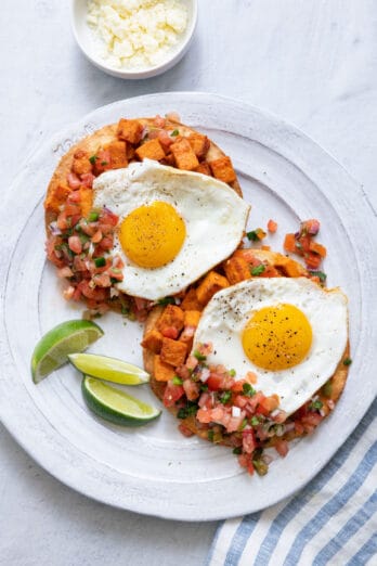 Large white plate with two huevos rancheros topped with sunny up eggs and garnished with lie wedges.