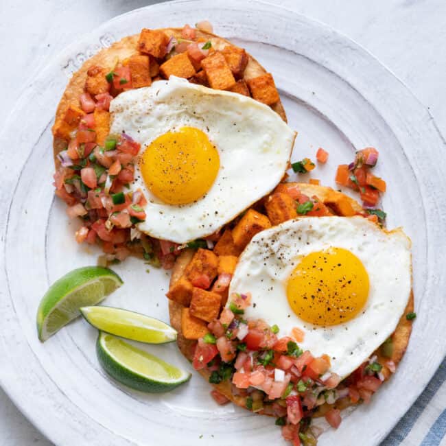 Large white plate with two huevos rancheros topped with sunny up eggs and garnished with lie wedges.