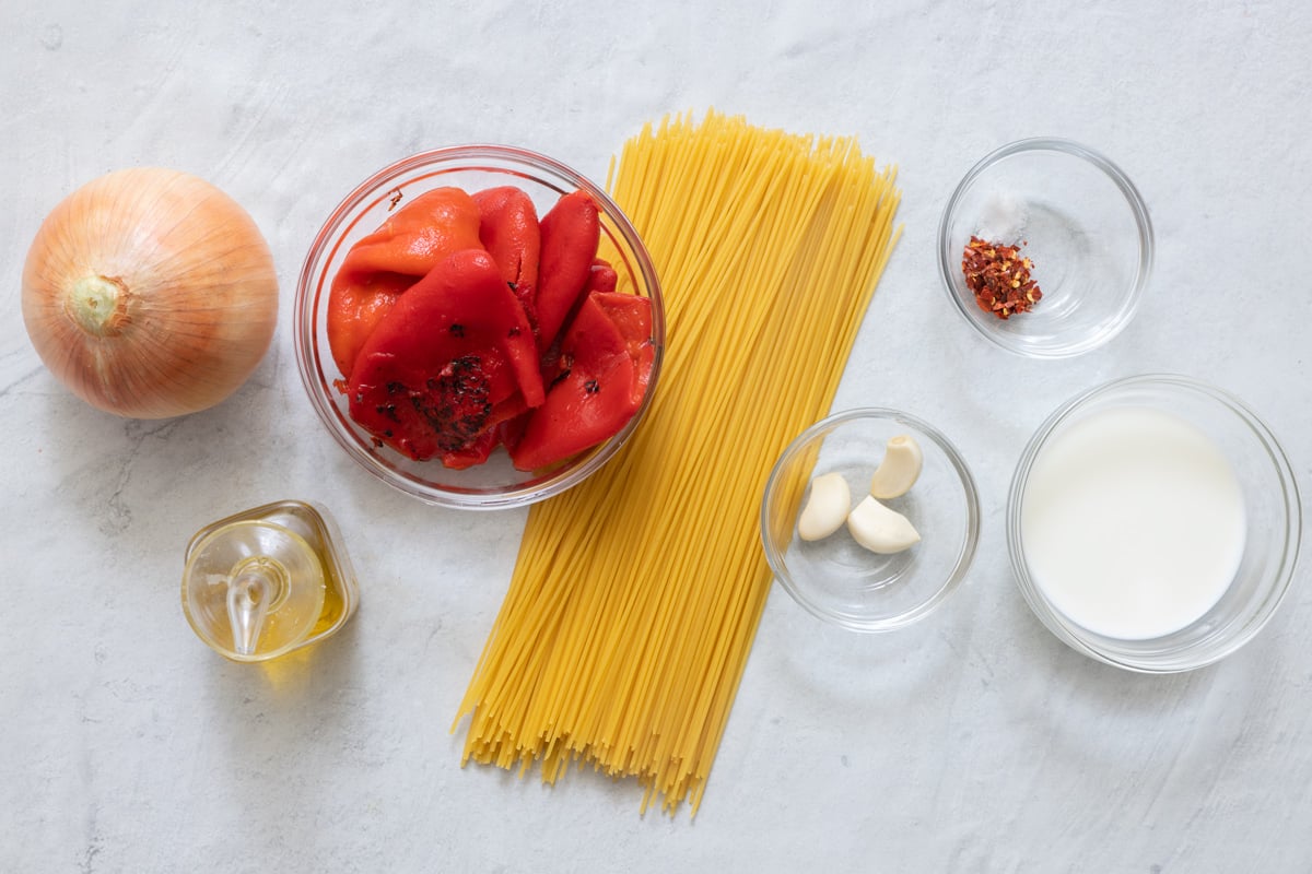 Ingredients for pasta sauce recipe before being prepped from left to right: whole onion, oil, roasted red peppers, dry noodles, garlic cloves, seasonings, and milk.