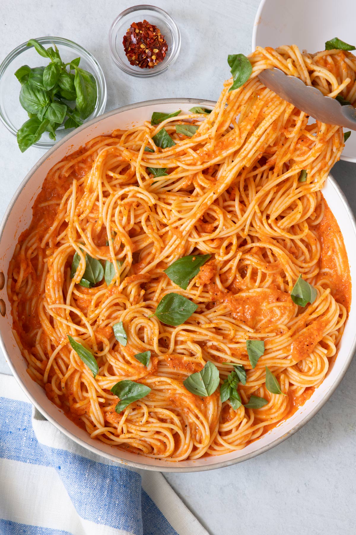 Tongs scooping pasta in a red sauce garnished with torn fresh basil from pot and a side of fresh basil and red pepper flakes in individual bowls.