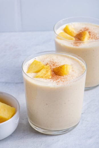 Two cups of mango pineapple smoothies with bowl of mangos on the side