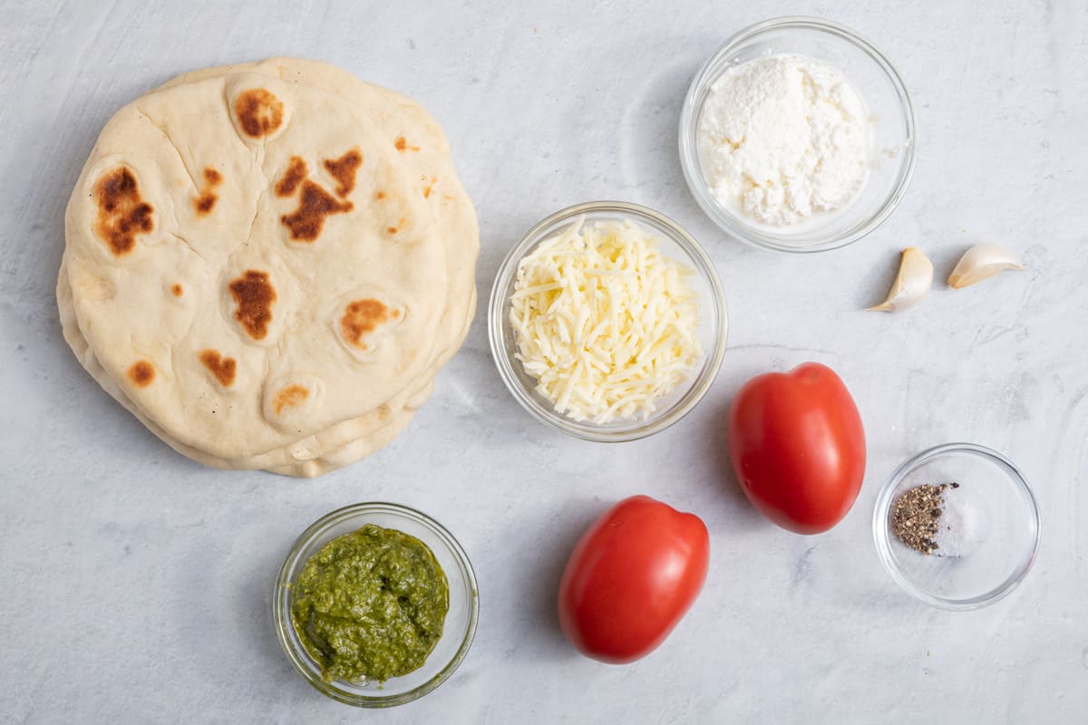 Ingredients for recipe on surface: flatbread, pesto, cheese, ricotta, roma tomatoes, seasoning, and 2 garlic cloves.