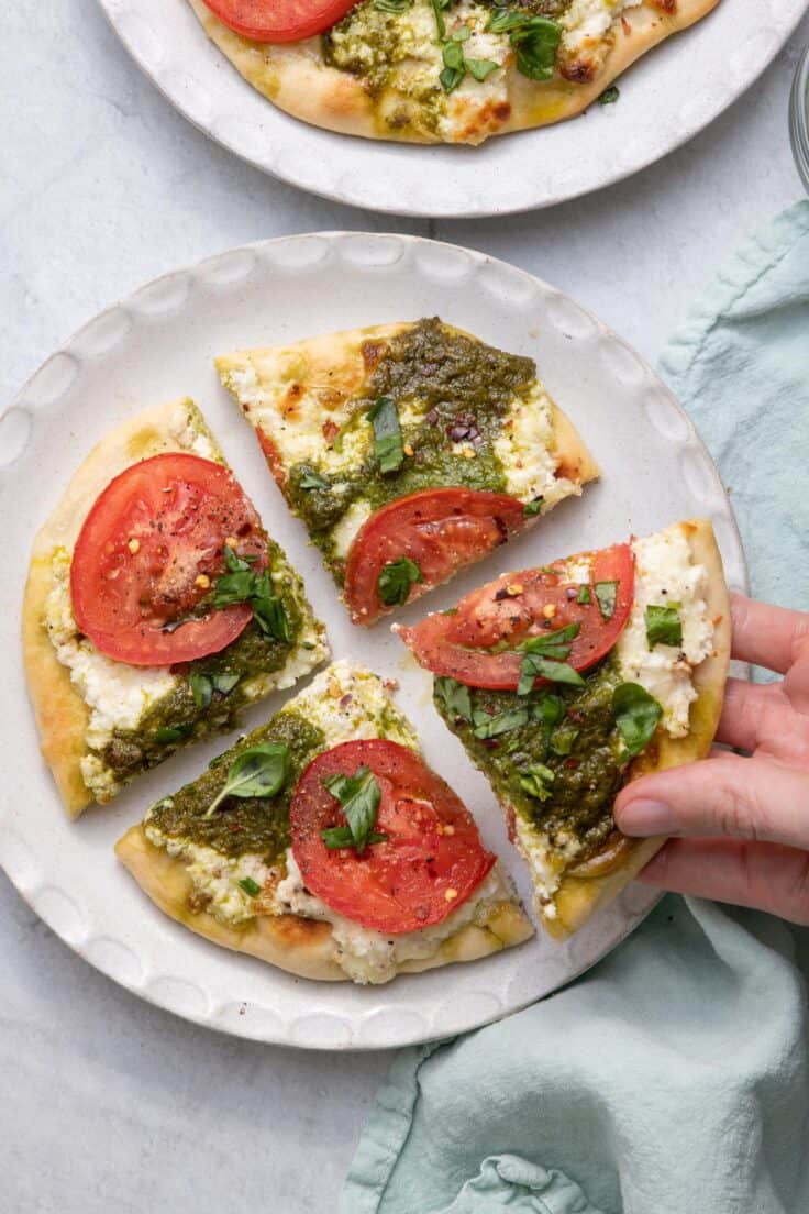 Pesto flatbread with sliced tomatoes on a plate cut into four slices with a hand grabbing a slice.
