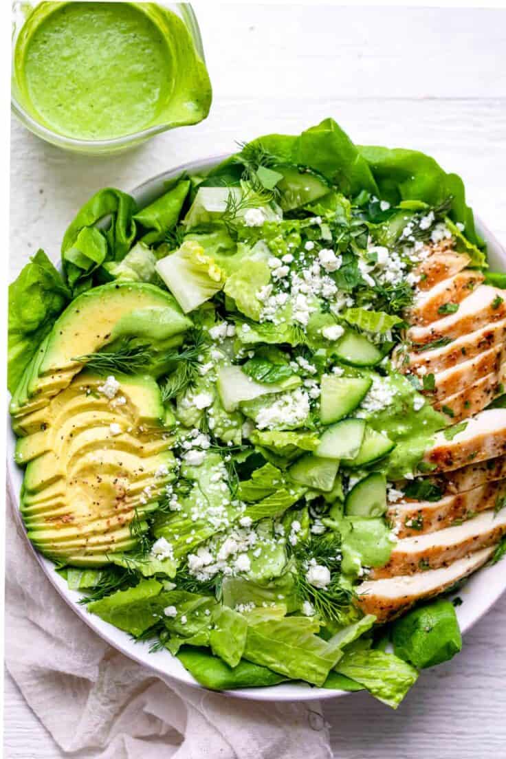 Sliced grilled chicken, sliced avocado, bed of greens in a white bowl plus green cilantro yogurt dressing in a clear jar