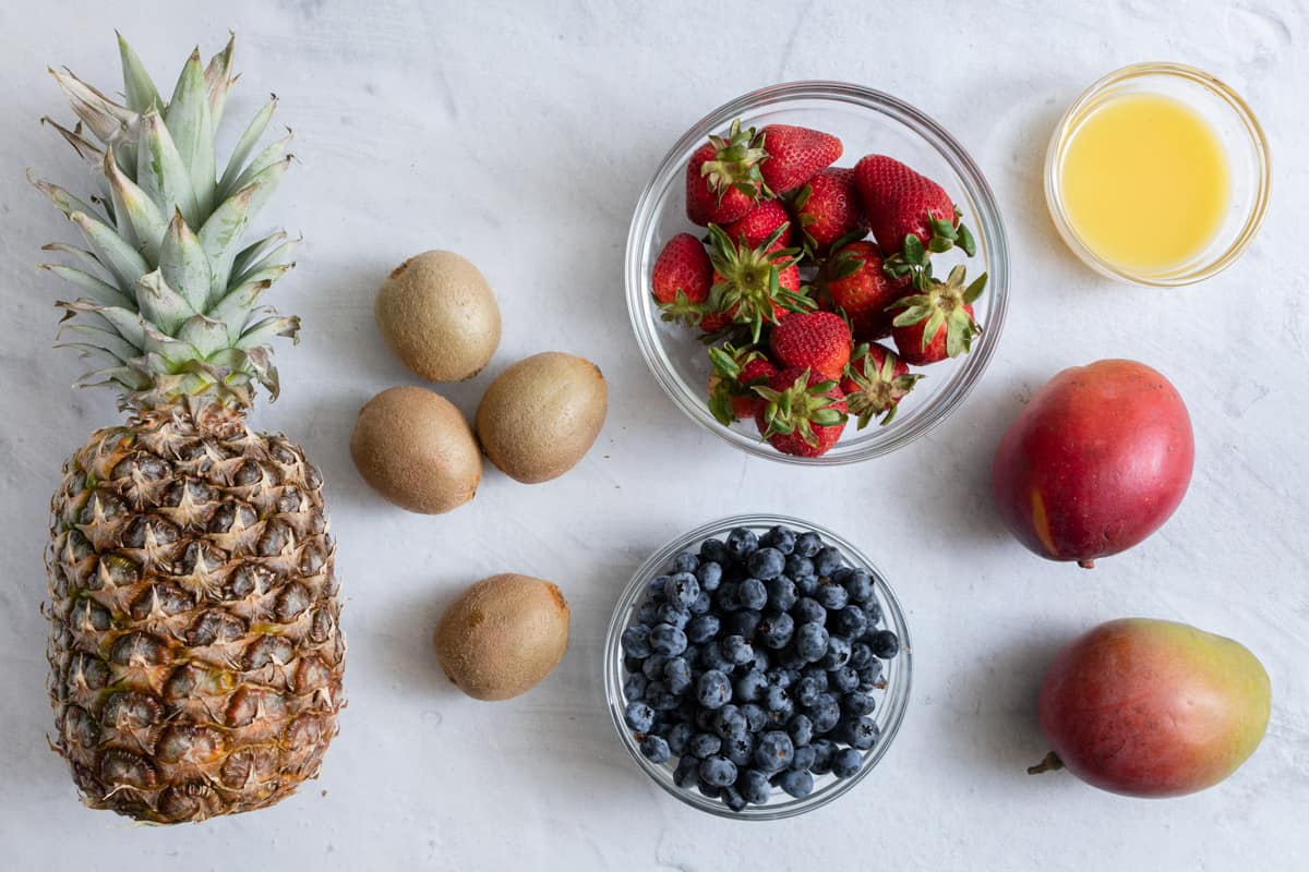 Ingredients for fruit salad: whole pineapple, kiwis, strawberries, blueberries, mangos, and orange juice.