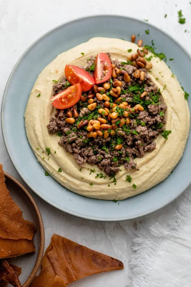 Blue plate with hummus, ground beef, pine nuts, parsley and tomatoes, with side of fried pita chips