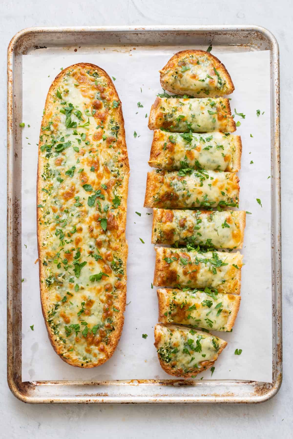Two baked garlic cheese bread on parchment paper line baking sheet with one bread cut into slices.
