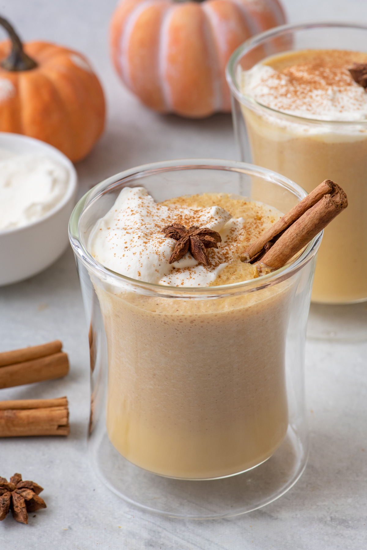 Glass of pumpkin latte with cinnamon sticks and whipped topping and another glass of shot in background.