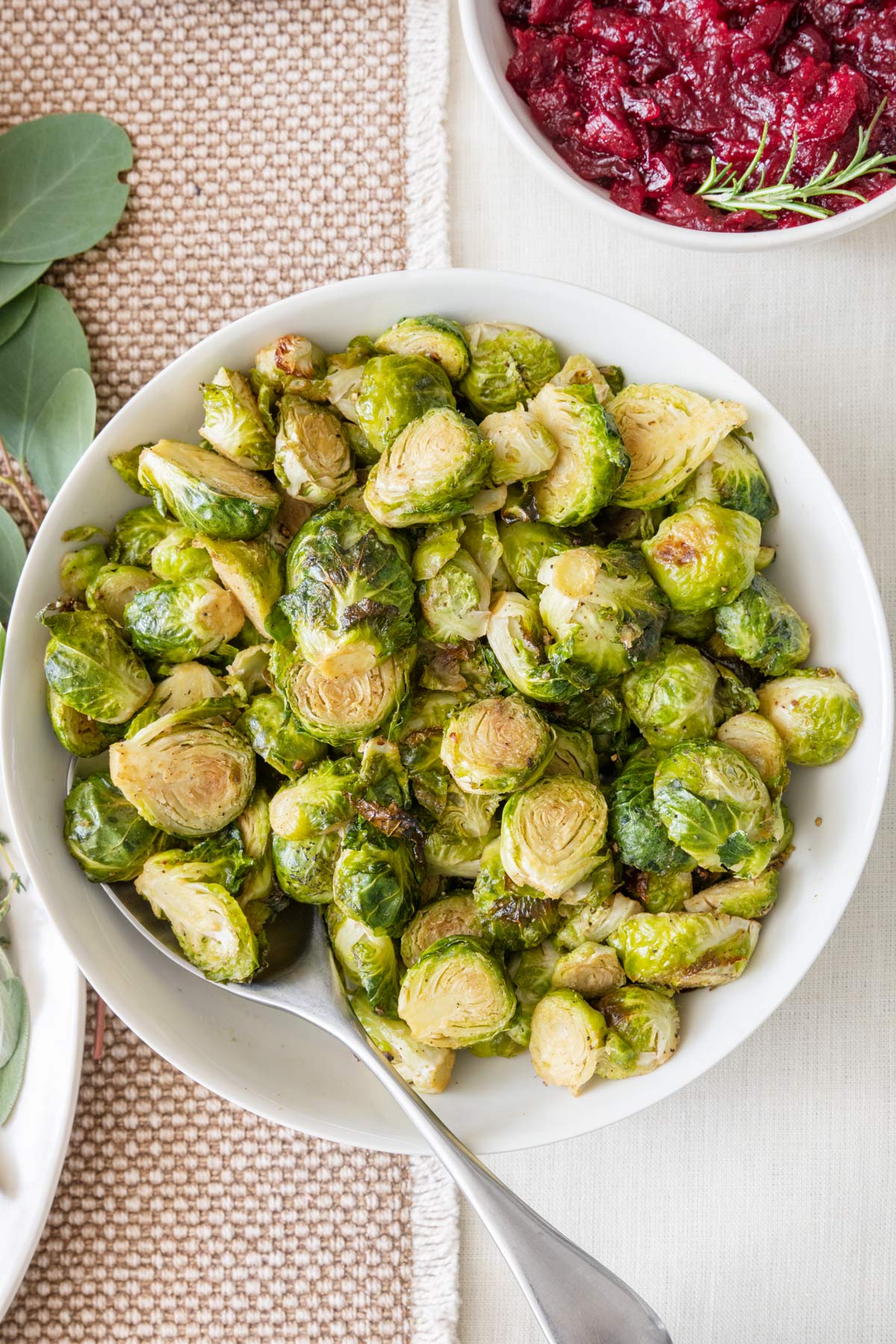 Roasted brussel sprout halves in a serving bowl with spoon inserted inside and a small dish of cranberry sauce nearby.