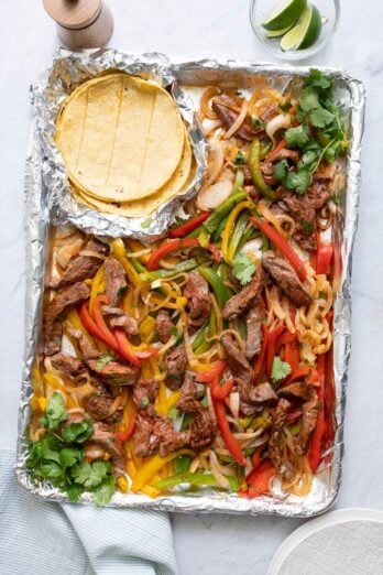Sheet pan with steak fajita ingredients tossed together, garnished with fresh cilantro with a side of corn tortilla shells in an open foil pack.