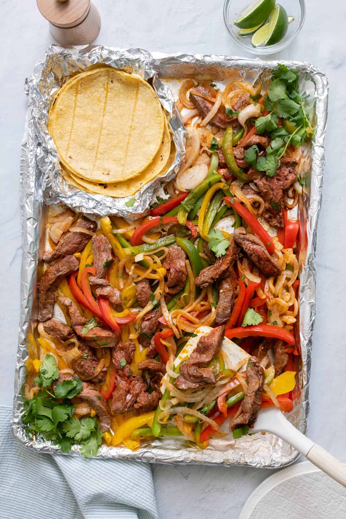 Spatula scooping a serving of steak fajitas from a sheet pan with a side of corn tortilla shells and lime wedges.