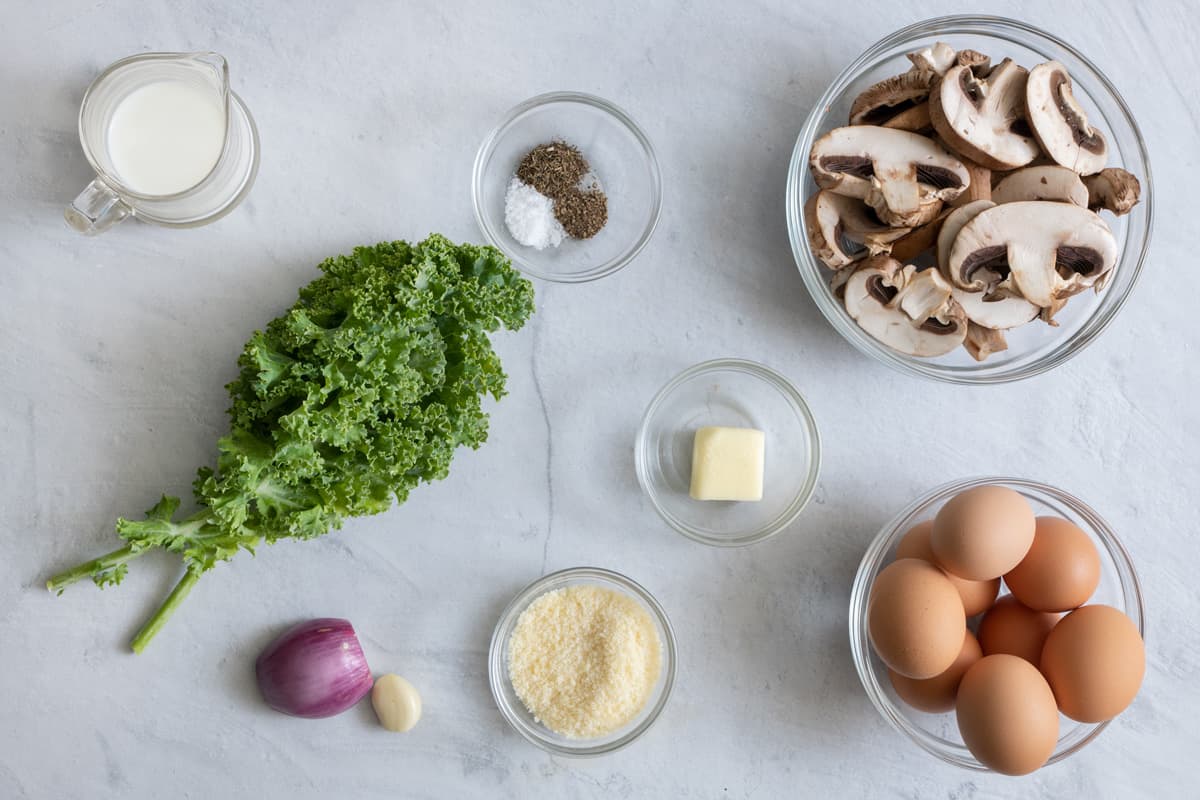 Ingredients for recipe before being prepped: milk, kale, shallot, garlic clove, parmesan, butter, spices, mushrooms, and shelled eggs.
