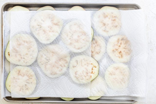 Eggplant slices on a baking dish with salt and paper towels over them to drain out the bitter liquid