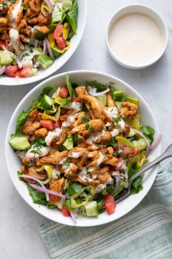 Two large bowls of chicken shawarma salad with tahini sauce drizzled on top and served on the side
