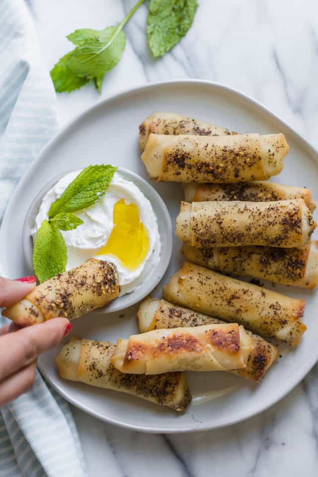 Plate of zaatar spring rolls with one spring roll being dipped into labneh yogurt dip with mint leaves.