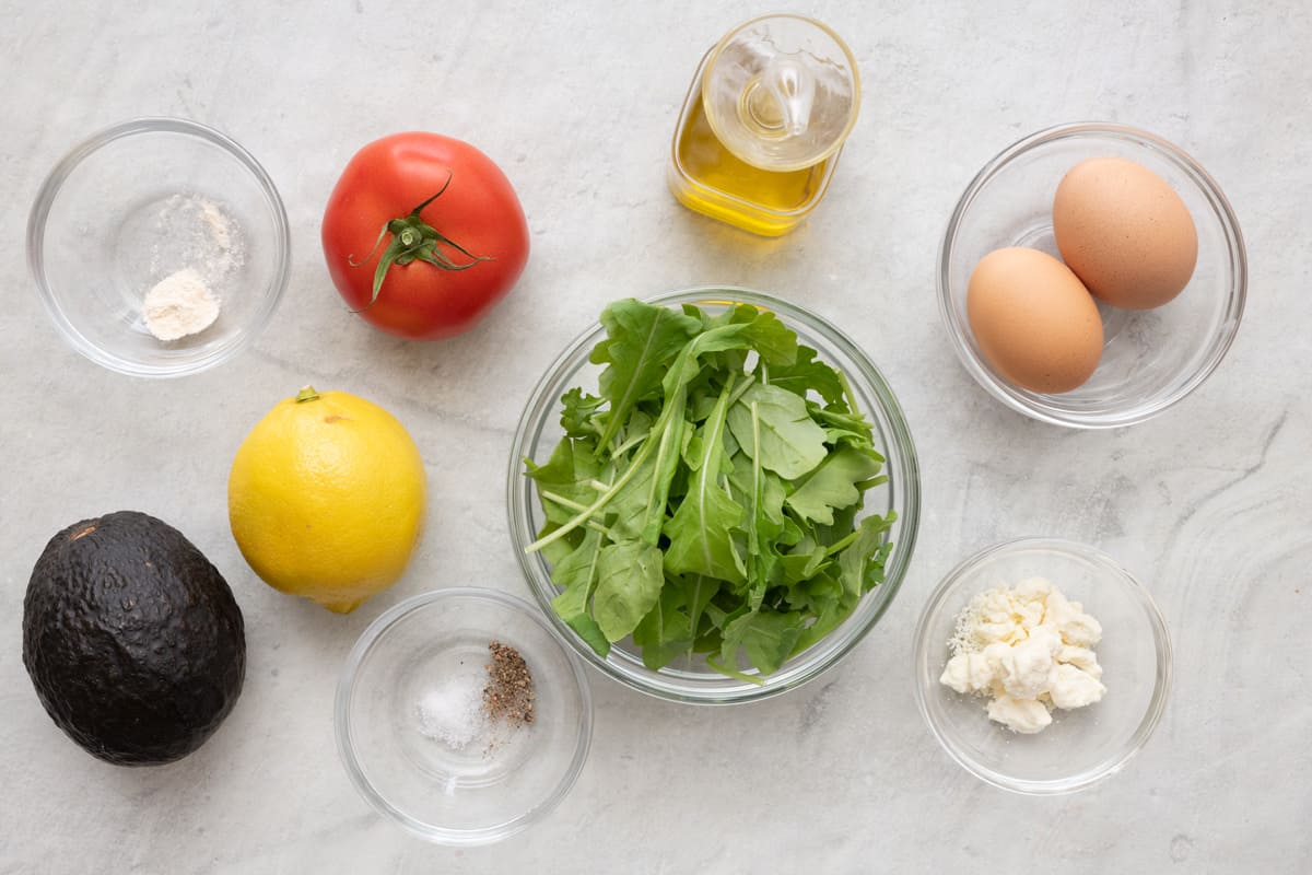 Ingredients for recipe in individual bowls and before prepped: flour, whole tomato, lemon, and avocado, salt and pepper, arugula, oil, 2 eggs, and crumbled feta cheese.