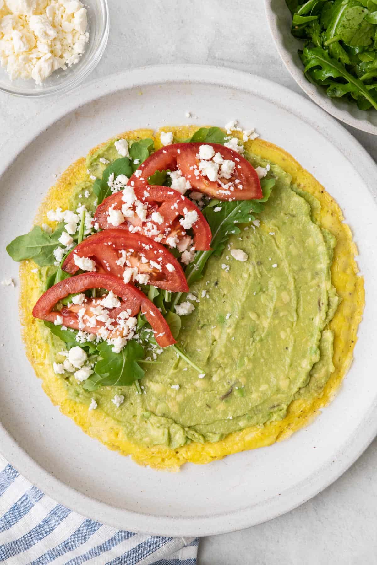 Egg wrap shell on plate with avocado mash spread on and then half topped with arugula, sliced tomato halves, and feta cheese.