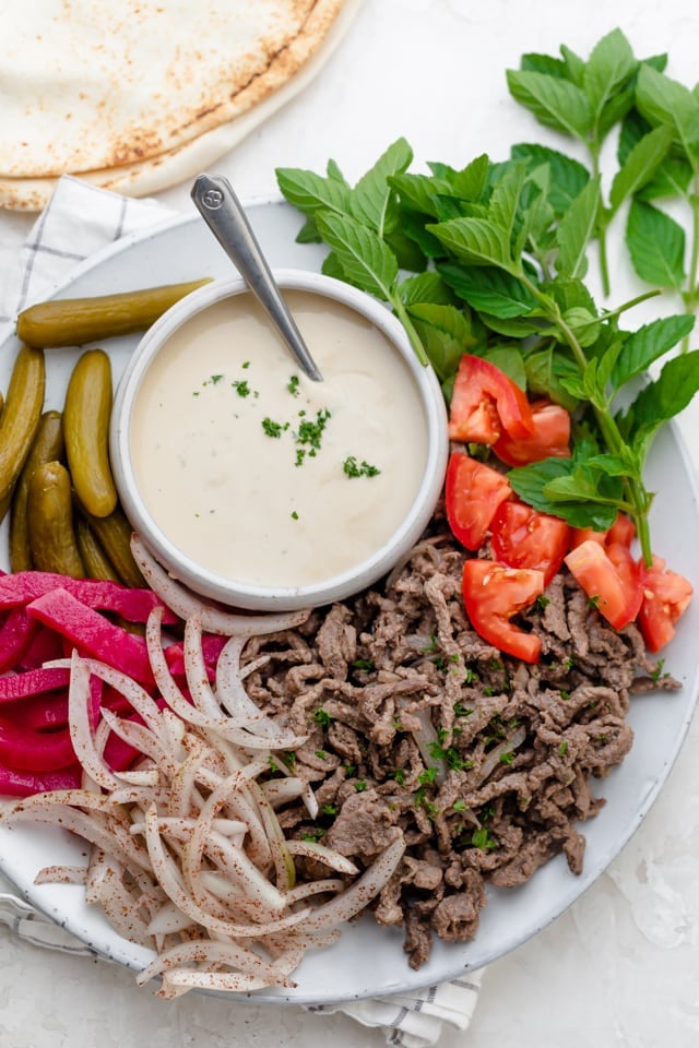 Cooked beef shawarma on a round plate, surrounded by all the accompaniments such as tomatoes, parsley, onions, pickle and pickled turnips