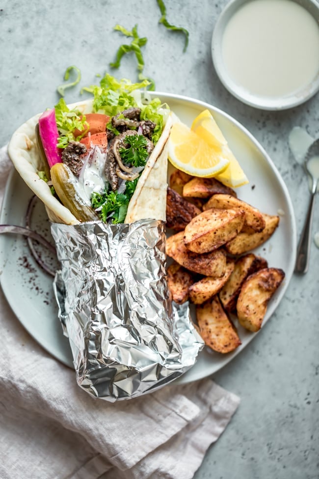 Beef shawarma sandwich rolled up in a thick pita with the bottom half of the wrap covered in aluminum foil sitting on a plate, alongside baked potato wedges and lemon wedges.