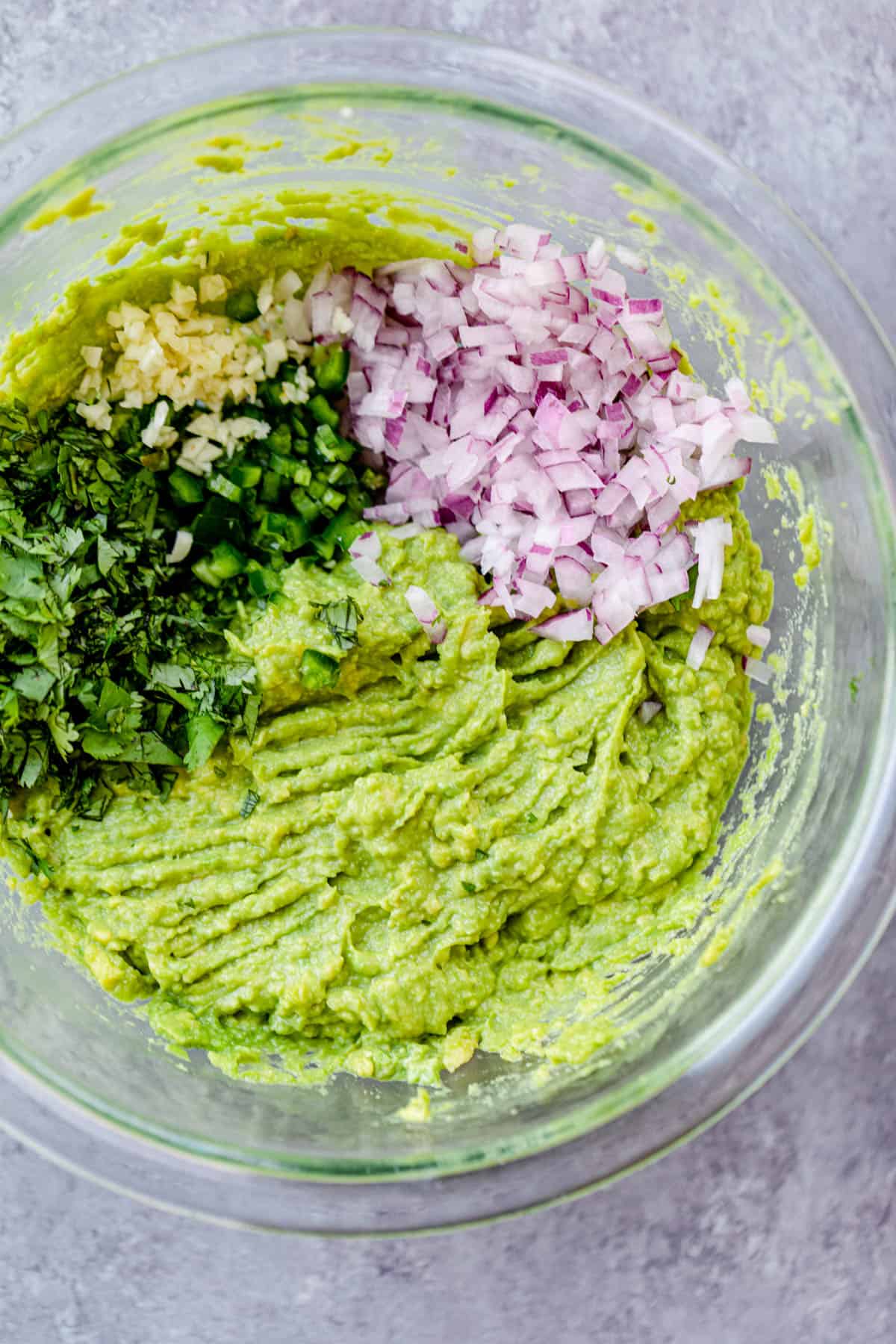 Big clear bowl with mashed avocados. On top of the mashed avocados are chopped red onions, chopped cilantro and chopped jalapeno