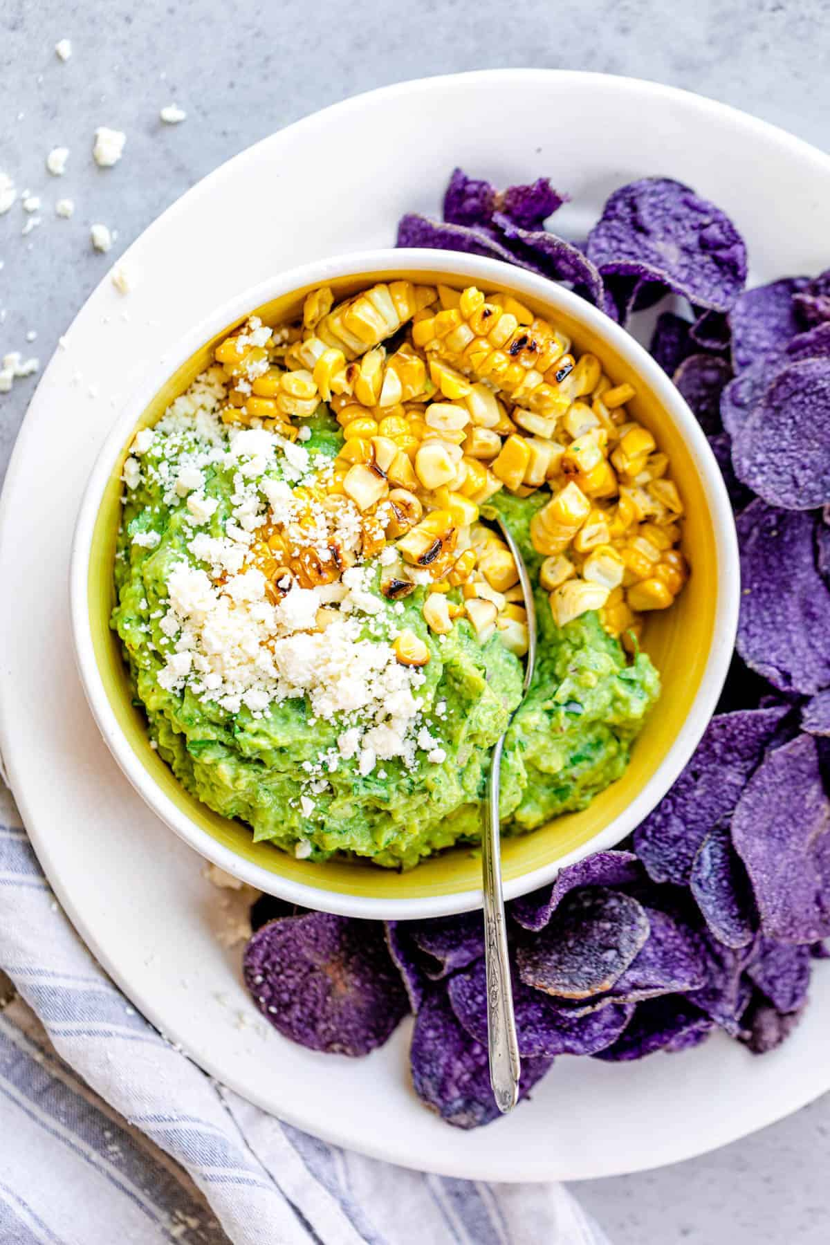 Bowl of the finished product - simple guacamole with grilled corn, cojita cheese. Around the bowl of guacamole is a round plate with purple potato chips