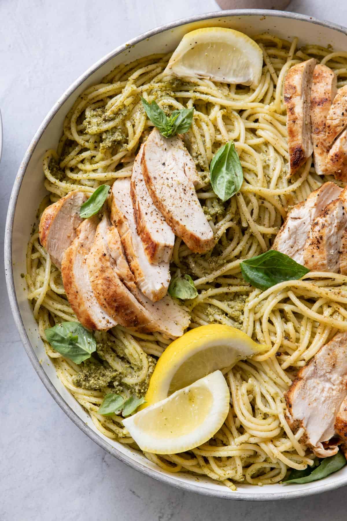 Close up of finished dish of pasta with pesto, sliced chicken in pot garnished with basil and lemon wedges.