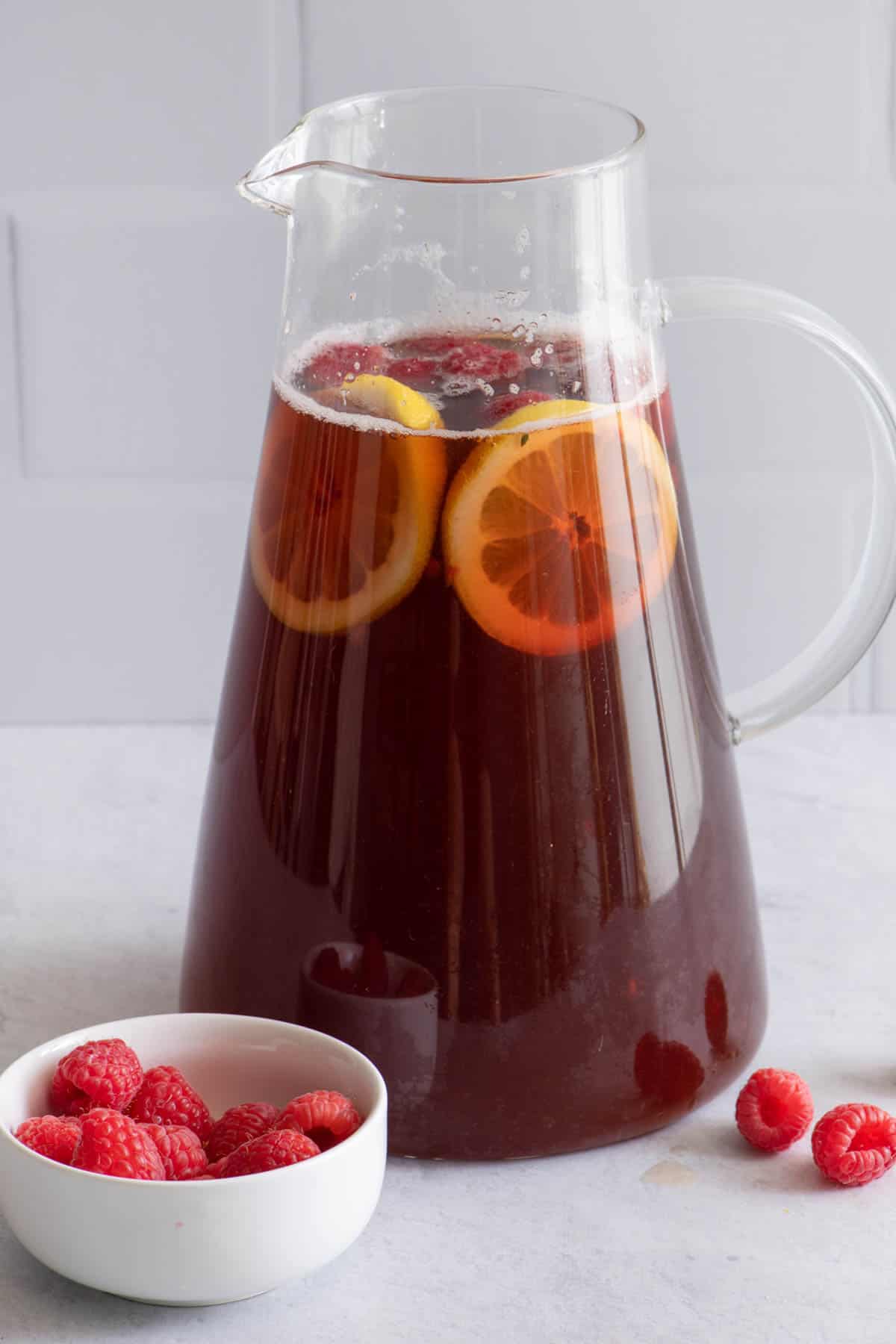 Large pitcher of raspberry tea with a halved lemon inside and a small bowl of fresh raspberries.