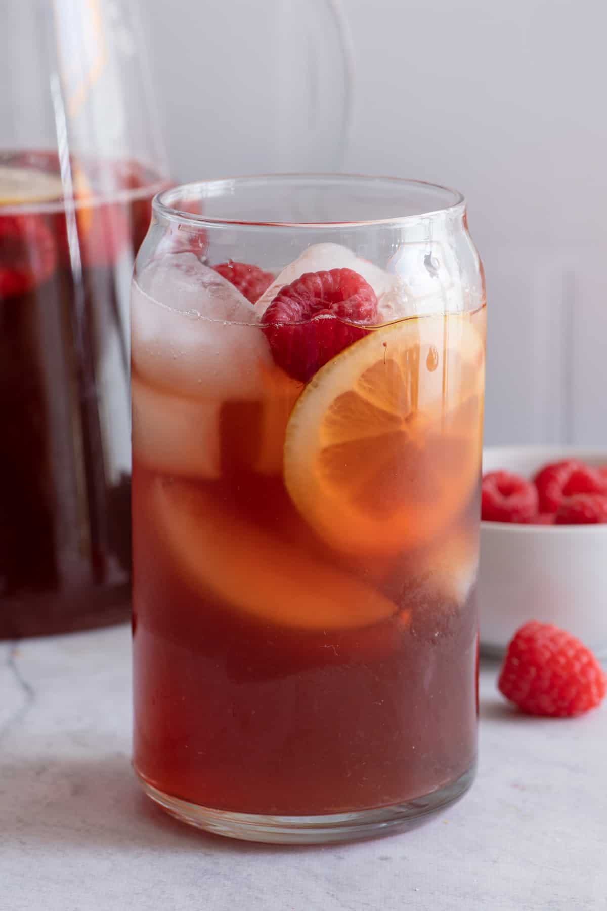 Large glass of raspberry iced tea with fresh cut lemons and raspberries and the pitcher of tea sitting behind.