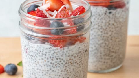 2 jars of chia seed pudding with chopped strawberries, blueberries, and just a few shreds of coconut on top with more fruit around jars.