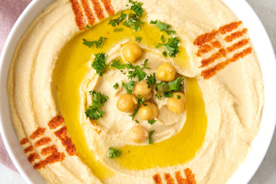 Bowl of authentic Lebanese hummus topped with olive oil, a few chickpeas, fresh chopped parsley, and a decorative lines made with paprike. Oil and pita nearby.