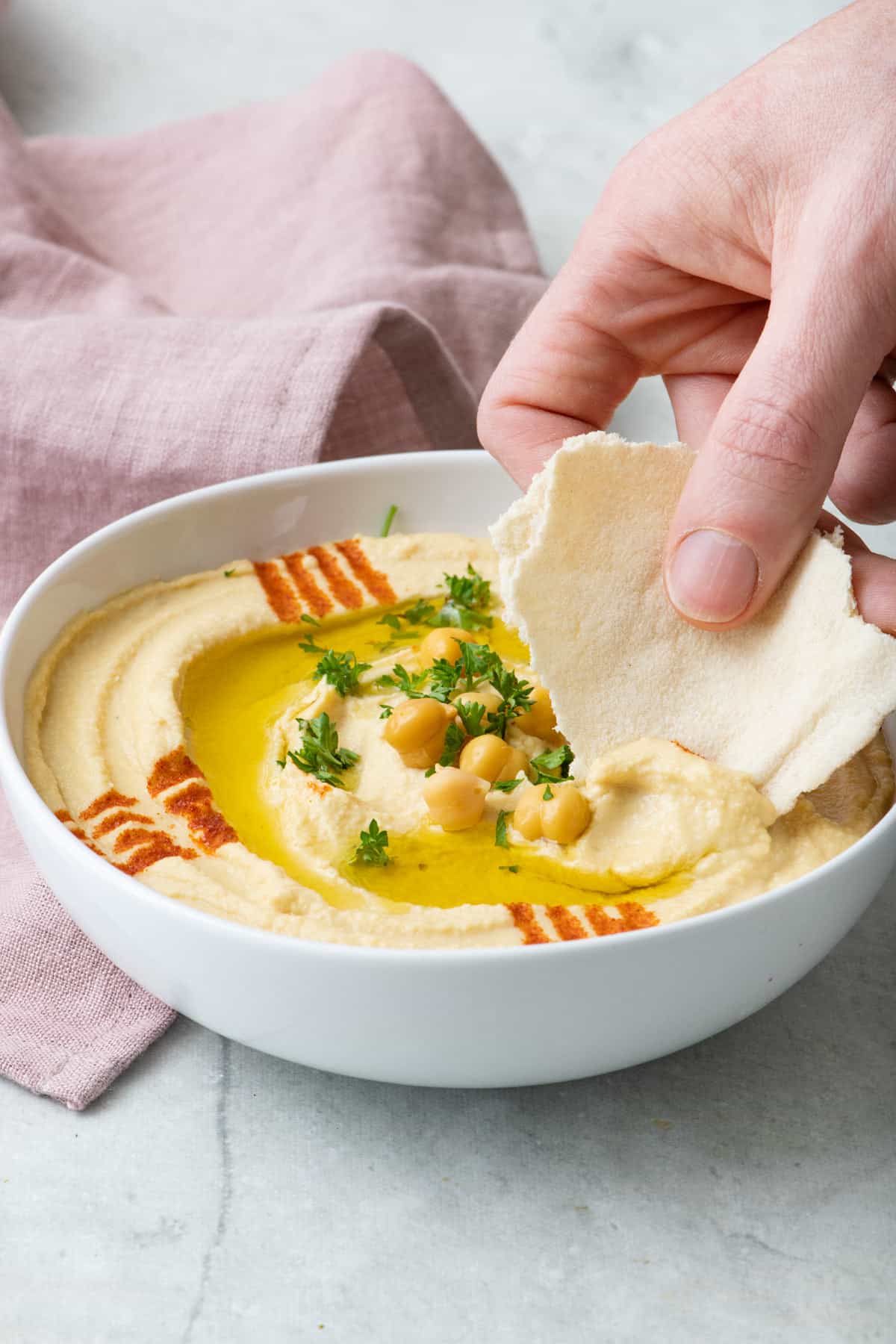 Final plated dish showing hummus with olive oil and garnished with paprika and parsley and an thin torn pita dipping into it.