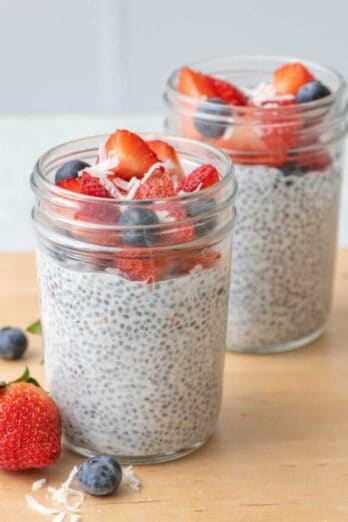 2 jars of chia seed pudding with chopped strawberries, blueberries, and just a few shreds of coconut on top with more fruit around jars.