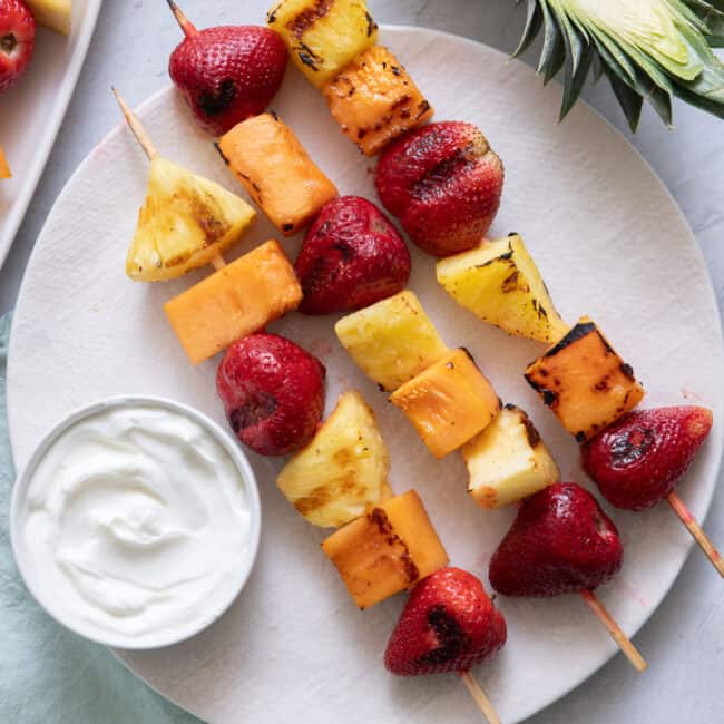 3 grilled fruit kabobs of cantaloupe, pineapples, and strawberries, with a side of yogurt for dipping.