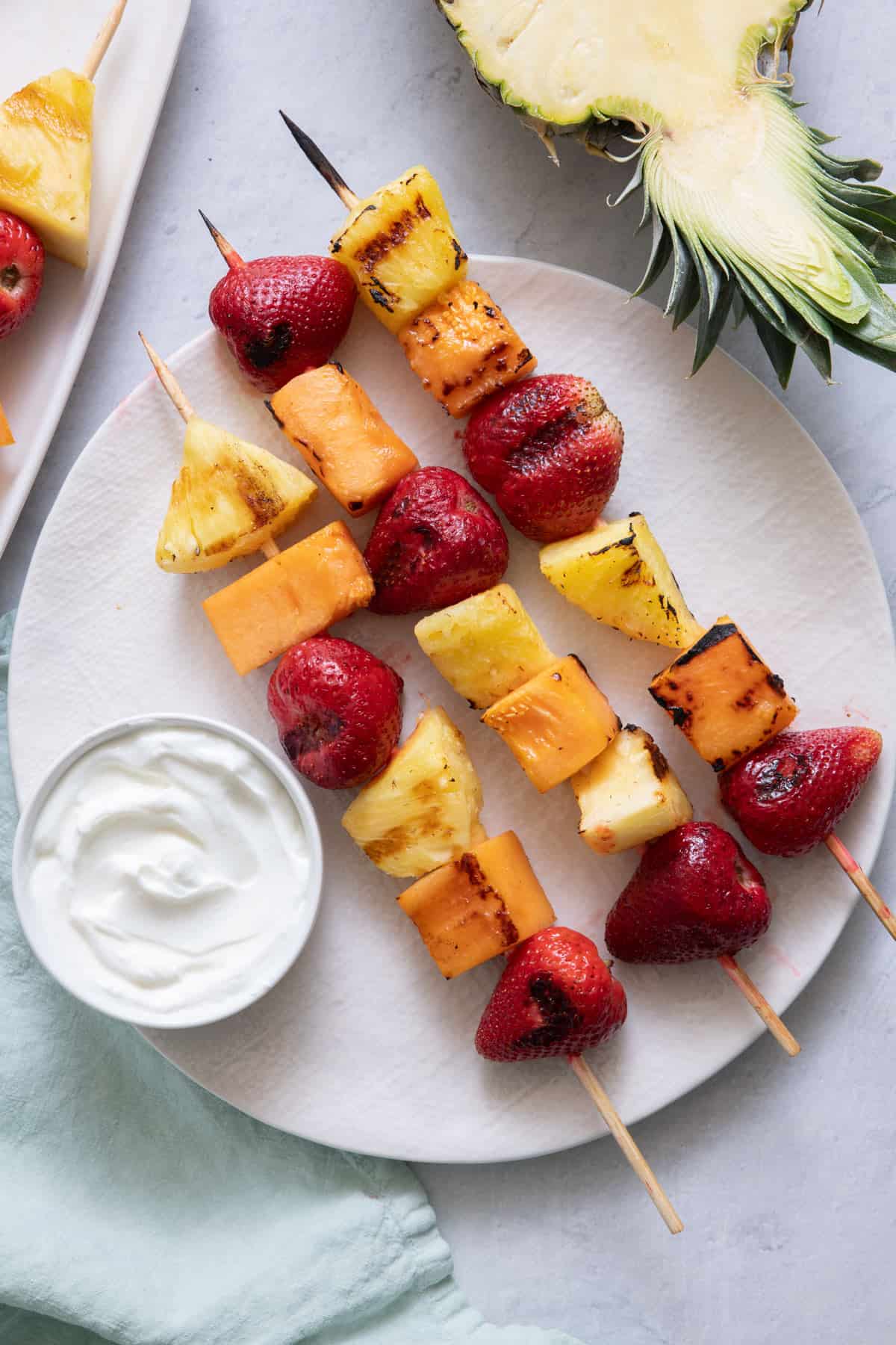 3 grilled fruit kabobs of cantaloupe, pineapples, and strawberries, with a side of yogurt for dipping.